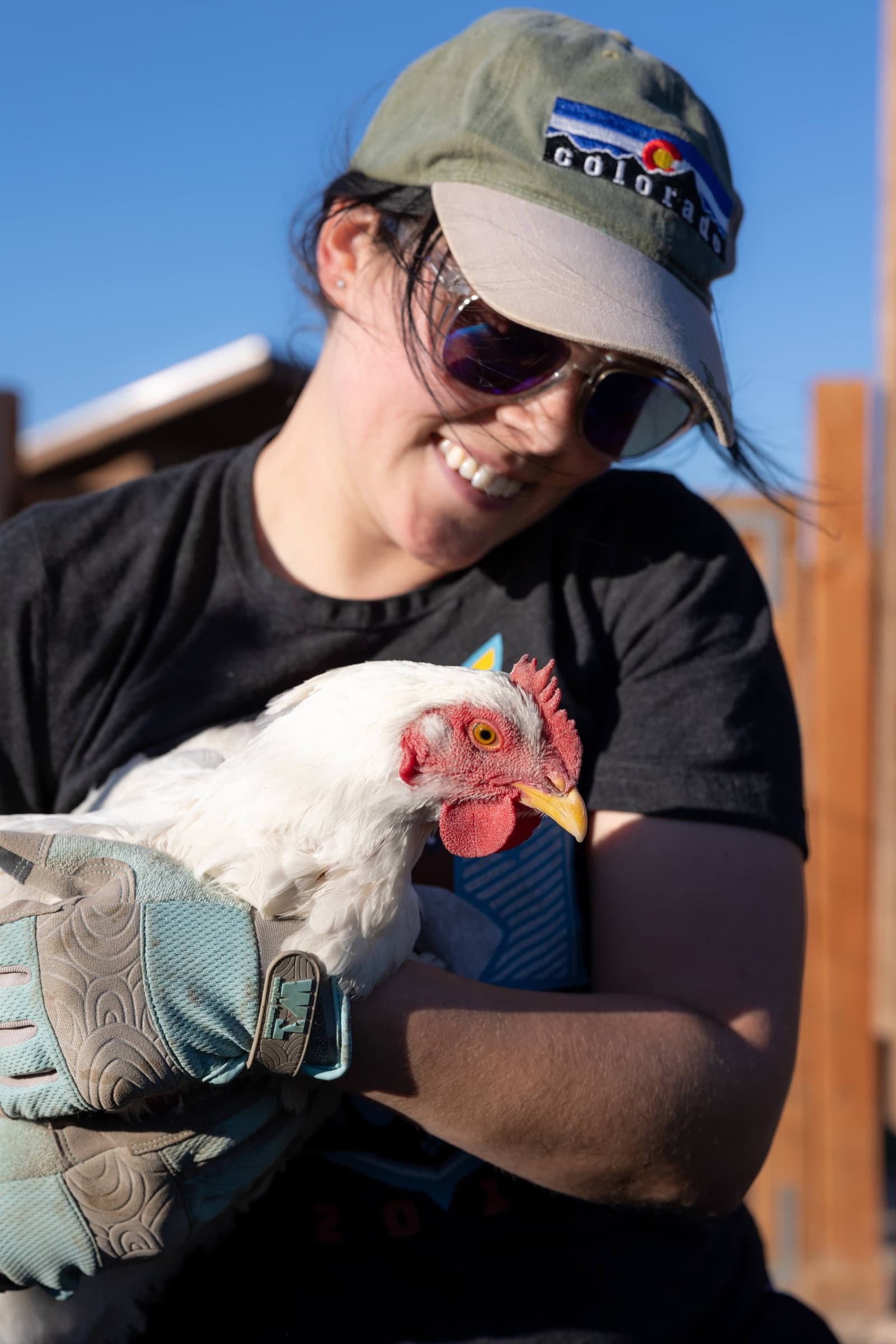 A woman in a cap and sunglasses smiling down at the white hen she is holding in her arms, blue sky behind them